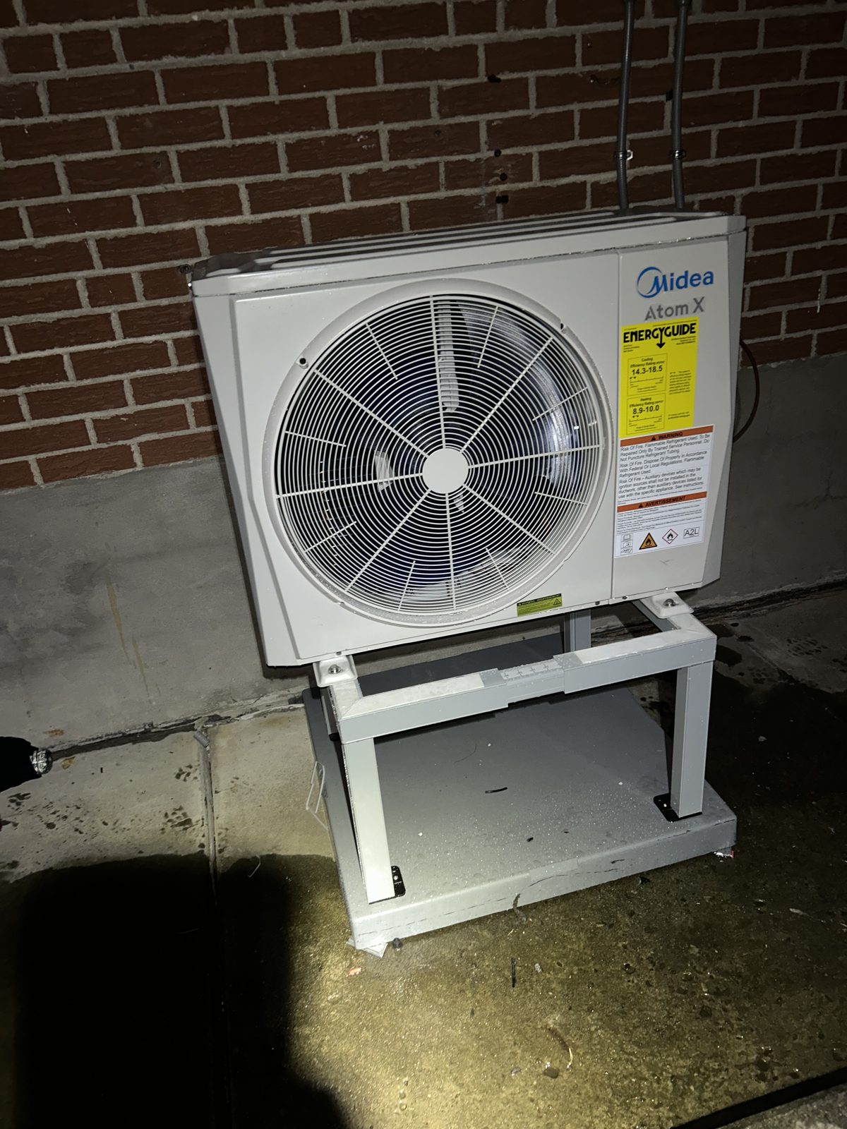 Midea Atom X outdoor condenser mounted on a raised steel bracket against a brick wall